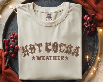 May include: Cream-colored t-shirt with the words "HOT COCOA WEATHER" in brown, vintage-style lettering. The shirt is folded neatly on a dark metal tray, with sprigs of red berries and string lights in the background.