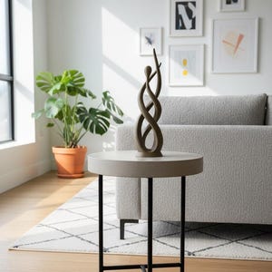 Pode incluir: Uma sala de estar moderna com uma pequena mesa lateral redonda. A mesa tem um tampo cinza claro e pernas de metal preto. Uma escultura abstrata marrom está em cima. Uma planta Monstera em vaso e obras de arte emolduradas estão ao fundo.