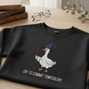 May include: Black sweatshirt with a cartoon goose wearing a wizard hat and holding a staff. The goose is white with orange feet and beak. The text "OFF TO COMMIT TOMFOOLERY" is printed below the goose in white.