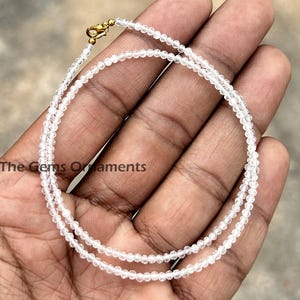 Könnte beinhalten: Zarte weiße Perlenkette mit goldenem Verschluss. Die Halskette besteht aus kleinen, runden Perlen und wird auf einer Hand präsentiert. Der Text "The Gems Ornaments" ist sichtbar.