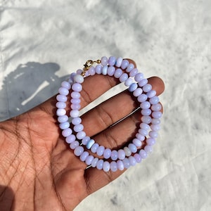 May include: A delicate necklace composed of small, light purple beads. The beads are strung together, forming a double strand. The necklace is secured with a gold-colored clasp. The beads have a smooth, rounded shape, and the overall design is simple and elegant.