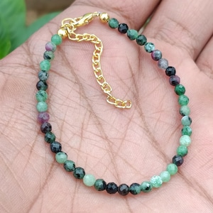 Può includere: Un braccialetto di perline con piccole pietre rotonde sfaccettate nei toni del verde, nero e magenta. Il braccialetto presenta una chiusura a moschettone in metallo dorato e una catenella di estensione regolabile, creando un accessorio delicato.