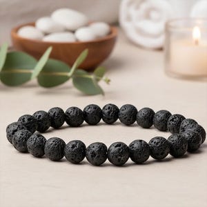 Może przedstawiać: Czarna bransoletka z kamieni lawowych z okrągłymi, teksturowanymi koralikami. Bransoletka jest prezentowana na neutralnej powierzchni, z drewnianą miską z białymi kamieniami, zielenią i zapaloną świecą w tle.