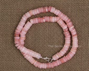 May include: A pink beaded necklace with a silver clasp. The necklace is made of small, flat, round beads in varying shades of pink. The beads are strung together to form a double strand necklace. The necklace is displayed on a brown textured surface.