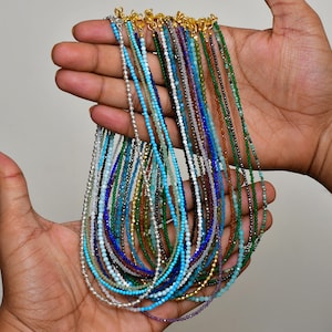 Könnte beinhalten: Eine Sammlung mehrerer zarter Perlenketten mit kleinen, facettierten Edelsteinen in verschiedenen Farben wie Türkis, Blau, Grün, Violett und Metallic-Silber, jeweils versehen mit einem kleinen goldfarbenen Karabinerverschluss aus Metall.