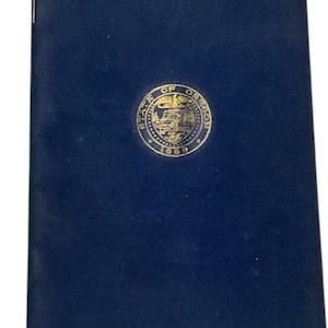 May include: A dark blue book with a gold emblem in the center. The emblem appears to be a seal with text around the perimeter. A gold-colored clasp is visible at the bottom edge of the book.