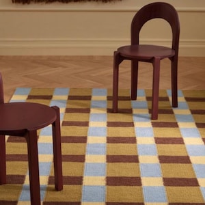 May include: Two burgundy-colored wooden chairs with rounded backs stand on a checkered rug. The rug features a pattern of brown, yellow, and light blue squares. A woven wall hanging is visible in the background.