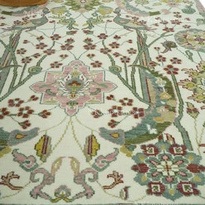 May include: Cream-coloured area rug with an intricate floral and bird design. The rug showcases a central medallion in pink, green, and grey, encircled by stylized flowers and birds. The border is a darker pink with a fringed edge.