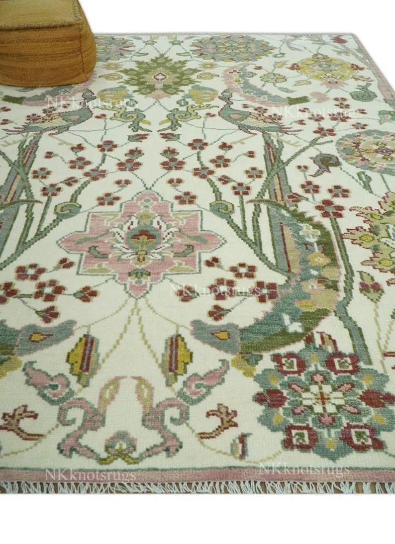 May include: Cream-coloured area rug with an intricate floral and bird design. The rug showcases a central medallion in pink, green, and grey, encircled by stylized flowers and birds. The border is a darker pink with a fringed edge.