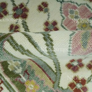 May include: Close-up of a rolled rug with a floral design. The rug has a cream background with repeating floral patterns in pink, burgundy, green, and yellow. The text "NKknotsrugs" is visible.