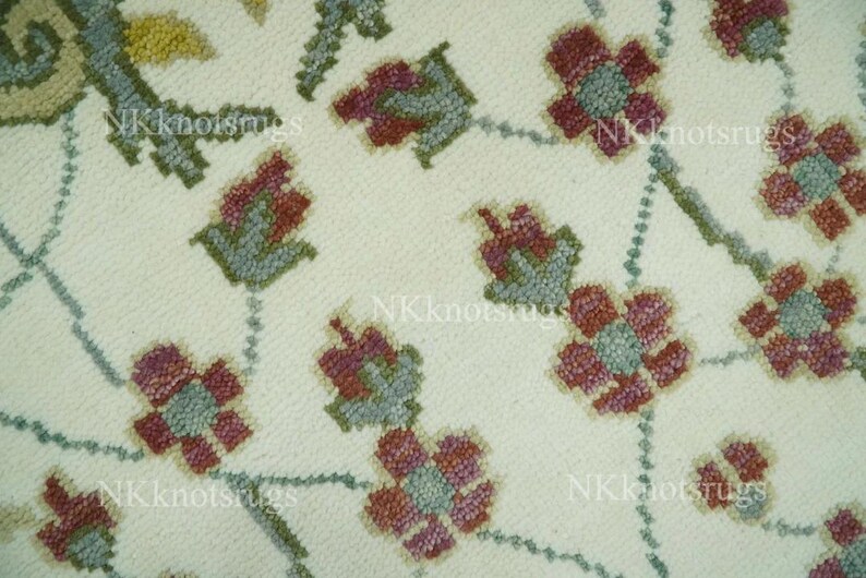 May include: Close-up of a cream-coloured rug with a floral pattern. The design features stylised flowers in shades of burgundy and teal, connected by a dotted vine pattern. The rug's texture is visible, suggesting a woven or embroidered construction.