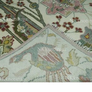 May include: Cream-coloured rug with intricate floral and bird patterns in shades of pink, green, and burgundy. The rug's corner is folded back, revealing the underside with a detailed bird design. The rug has a fringed edge.