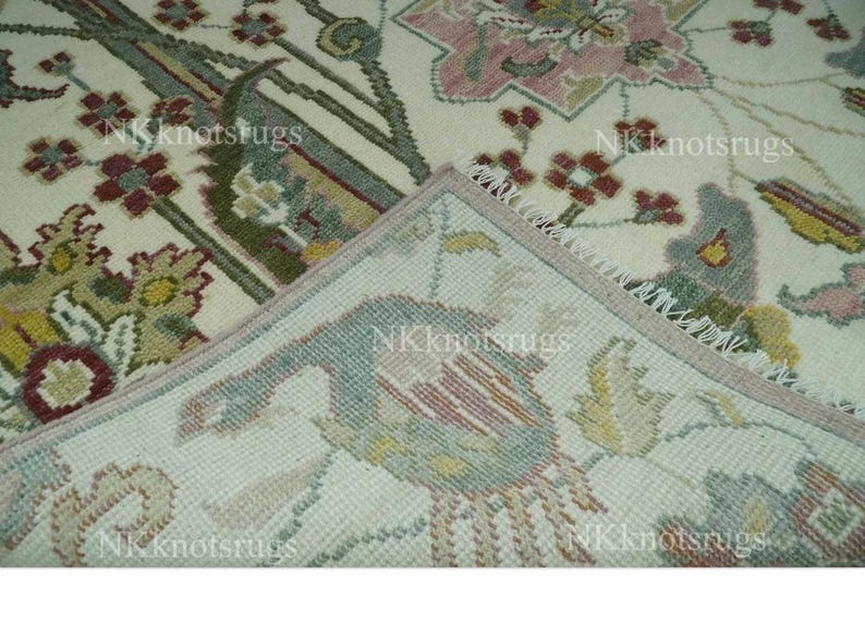 May include: Cream-coloured rug with intricate floral and bird patterns in shades of pink, green, and burgundy. The rug's corner is folded back, revealing the underside with a detailed bird design. The rug has a fringed edge.
