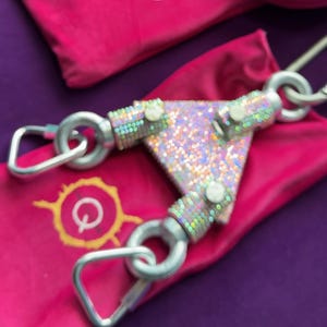May include: A triangular metal connector featuring a holographic, iridescent finish with three silver-toned carabiner clips attached to its corners. The device rests on a bright pink fabric pouch that displays a yellow circular logo with the letter Q.