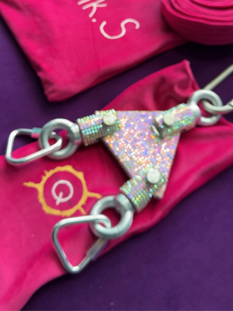 May include: A triangular metal connector featuring a holographic, iridescent finish with three silver-toned carabiner clips attached to its corners. The device rests on a bright pink fabric pouch that displays a yellow circular logo with the letter Q.
