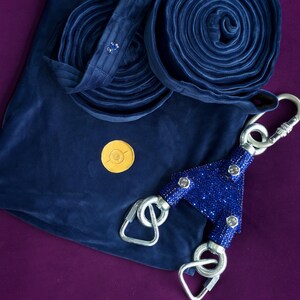 May include: A dark blue velvet aerial silk fabric folded into two circular coils, accompanied by a dark blue velvet storage bag, a small gold circular emblem, and a Y-shaped rigging plate covered in sparkling blue rhinestones with three steel carabiners.