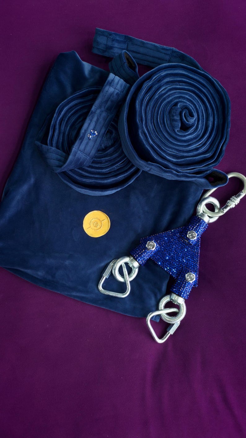May include: A dark blue velvet aerial silk fabric folded into two circular coils, accompanied by a dark blue velvet storage bag, a small gold circular emblem, and a Y-shaped rigging plate covered in sparkling blue rhinestones with three steel carabiners.