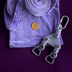 May include: A lavender velvet fabric accessory featuring two coiled straps, a circular gold-toned emblem, and a triangular metal connector piece encrusted with shimmering clear rhinestones, attached to three silver-toned metal swivel snap hooks.