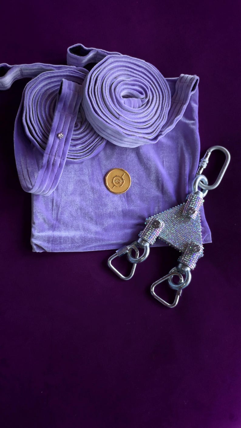 May include: A lavender velvet fabric accessory featuring two coiled straps, a circular gold-toned emblem, and a triangular metal connector piece encrusted with shimmering clear rhinestones, attached to three silver-toned metal swivel snap hooks.