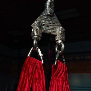 May include: A silver-colored metal aerial swivel plate with a triangular, textured surface, connected to a carabiner at the top and two lower attachment points. Two long, bright red fabric straps with a quilted texture hang from the metal hardware.