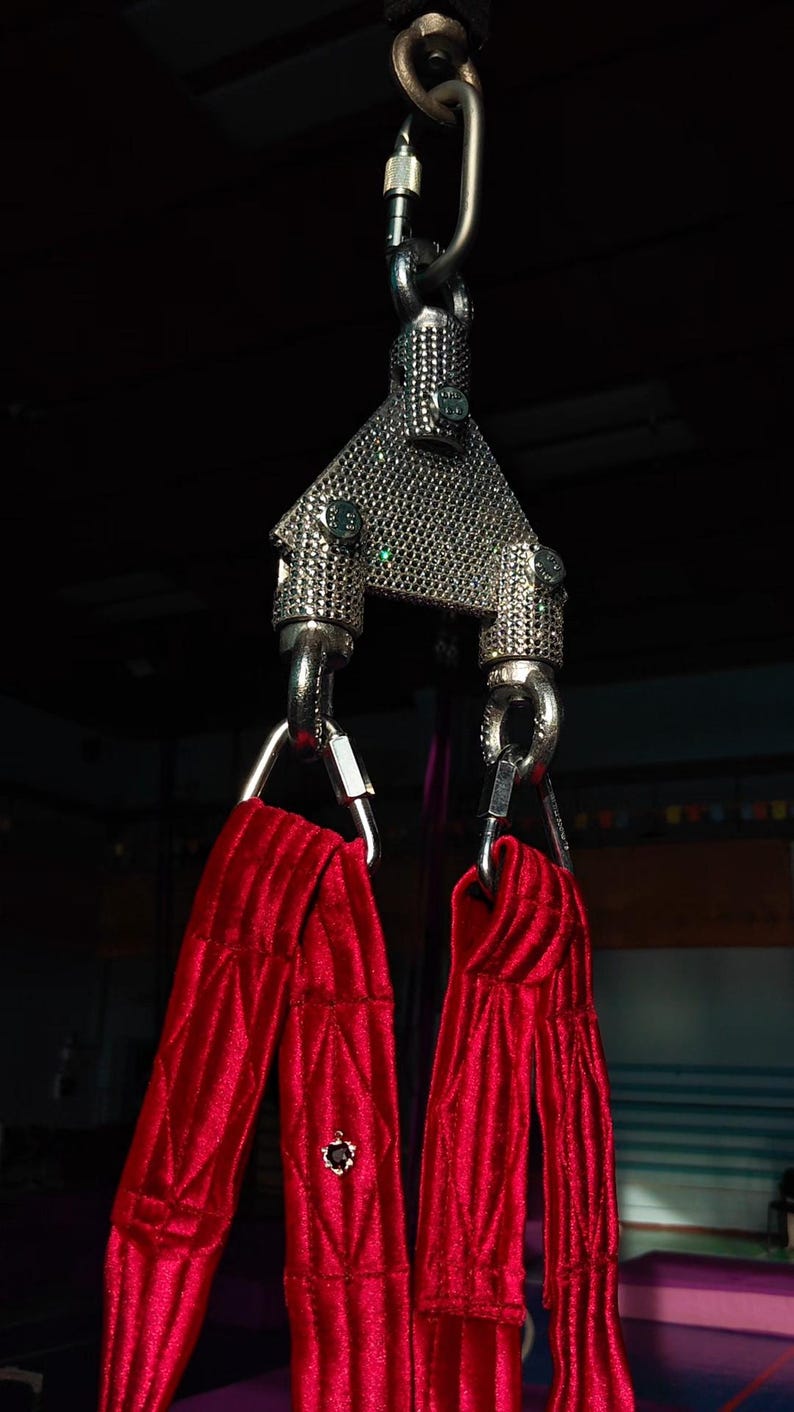 May include: A silver-colored metal aerial swivel plate with a triangular, textured surface, connected to a carabiner at the top and two lower attachment points. Two long, bright red fabric straps with a quilted texture hang from the metal hardware.