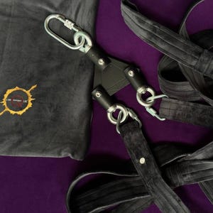 Puede incluir: Juego de bondage negro con mosquetones metálicos y una bolsa de almacenamiento de terciopelo negro. La bolsa tiene un diseño gráfico dorado y naranja. El equipo está sobre un fondo morado.