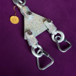 May include: A Y-shaped metal connector covered in iridescent rhinestones, featuring three silver-toned carabiners attached to the ends. A small gold-colored circular tag with an embossed symbol hangs from the top carabiner by a thin gold cord.