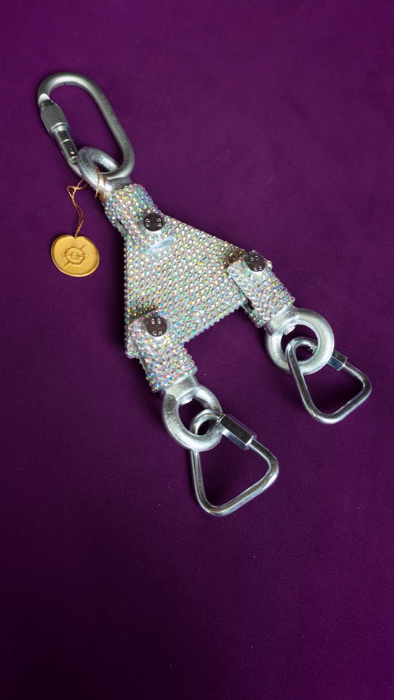May include: A Y-shaped metal connector covered in iridescent rhinestones, featuring three silver-toned carabiners attached to the ends. A small gold-colored circular tag with an embossed symbol hangs from the top carabiner by a thin gold cord.