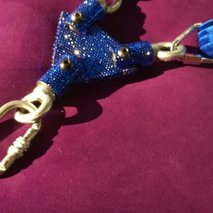 May include: A blue velvet strap assembly featuring a central triangular connector piece encrusted with small, shimmering blue rhinestones. The connector is attached to three silver-toned metal swivel snap hooks, providing a secure attachment point.