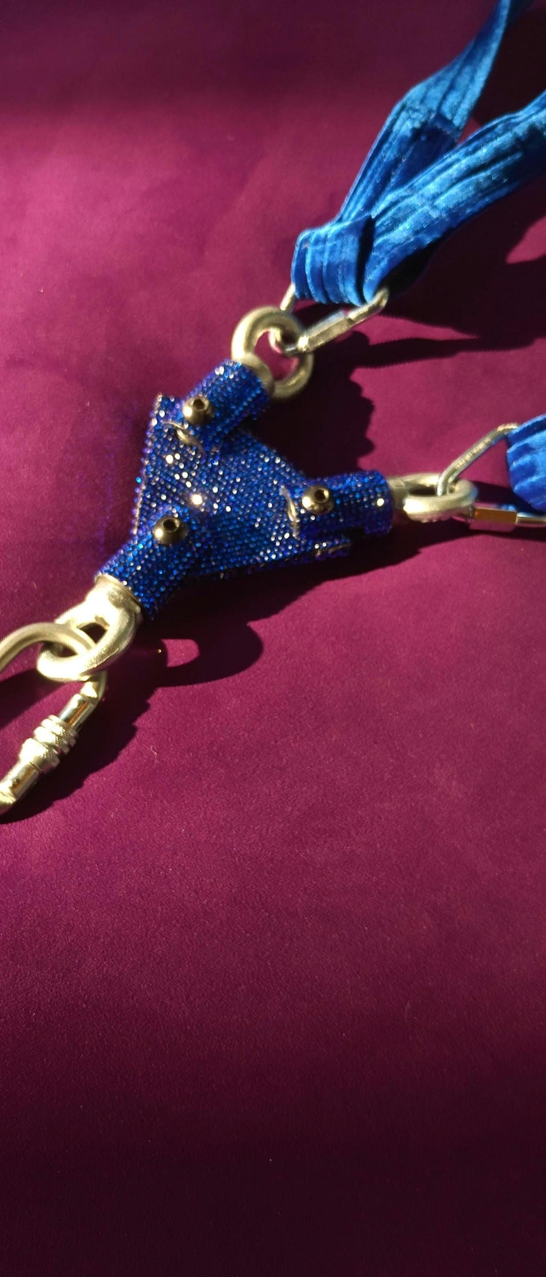 May include: A blue velvet strap assembly featuring a central triangular connector piece encrusted with small, shimmering blue rhinestones. The connector is attached to three silver-toned metal swivel snap hooks, providing a secure attachment point.