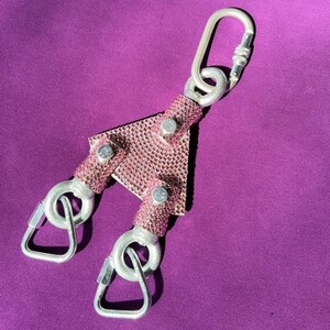 May include: A Y-shaped metal connector featuring a pink rhinestone-encrusted surface. The central triangular piece is attached to three silver-toned metal carabiners, each secured with a screw-gate locking mechanism for attachment purposes.