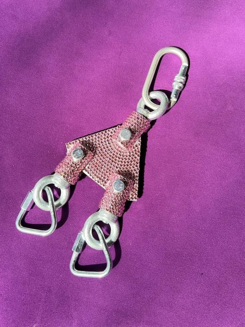 May include: A Y-shaped metal connector featuring a pink rhinestone-encrusted surface. The central triangular piece is attached to three silver-toned metal carabiners, each secured with a screw-gate locking mechanism for attachment purposes.