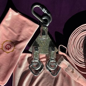 May include: A silver-toned metal swivel pulley encrusted with small clear rhinestones, featuring a large top carabiner and two smaller bottom carabiners. The pulley is surrounded by soft, light pink velvet fabric items with gold embroidery.
