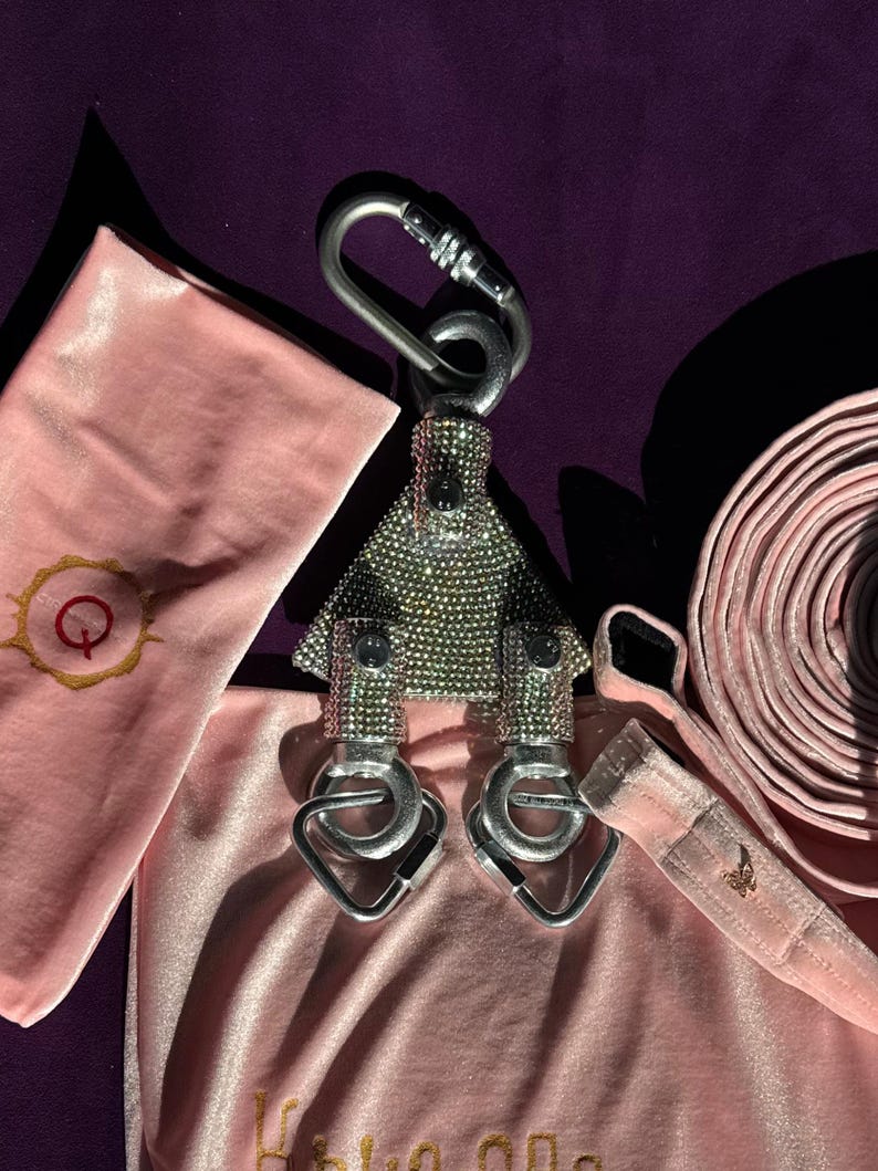 May include: A silver-toned metal swivel pulley encrusted with small clear rhinestones, featuring a large top carabiner and two smaller bottom carabiners. The pulley is surrounded by soft, light pink velvet fabric items with gold embroidery.