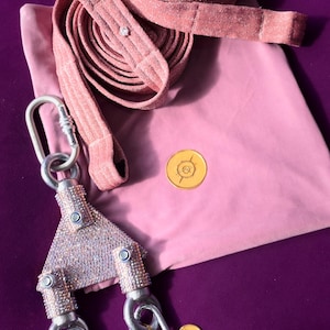 May include: A pink glittery long leash with a triangular metal connector piece covered in small clear crystals. The connector features two silver-toned carabiners at the bottom and one at the top. A pink fabric pouch with a gold circular logo is included.