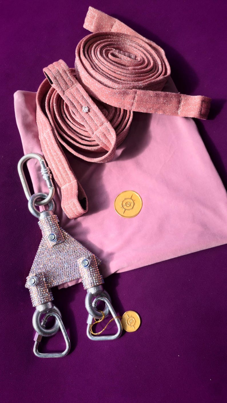 May include: A pink glittery long leash with a triangular metal connector piece covered in small clear crystals. The connector features two silver-toned carabiners at the bottom and one at the top. A pink fabric pouch with a gold circular logo is included.