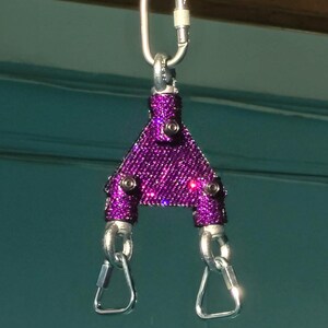 May include: A purple triangular spreader bar covered in small sparkling rhinestones, featuring three silver-toned metal carabiners attached to the top and bottom corners with visible black screws securing the metal frame structure.