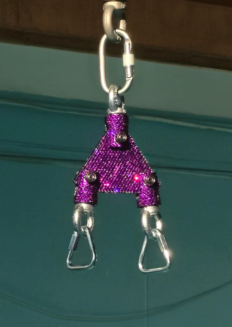 May include: A purple triangular spreader bar covered in small sparkling rhinestones, featuring three silver-toned metal carabiners attached to the top and bottom corners with visible black screws securing the metal frame structure.