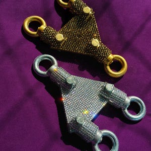 May include: Two triangular metal hardware connectors, one in gold tone and one in silver tone, both fully encrusted with small sparkling rhinestones. Each piece features three circular loops at the corners and three small metal bolts on the surface.