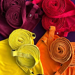 Puede incluir: Vista cenital de batas de terciopelo enrolladas en burdeos, rojo, amarillo y naranja. Cada bata tiene un cinturón a juego, algunos con detalles decorativos. Las batas están dispuestas sobre una superficie morada oscura.