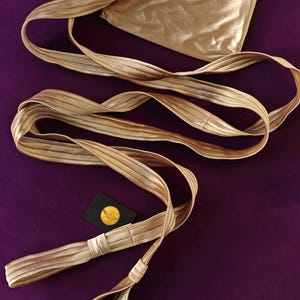 May include: A metallic gold fabric accessory featuring a long, pleated strap with stitched detailing and a square pouch. The pouch displays a circular emblem with a red letter Q inside a gold border. A small black tag with a gold circular logo is included.