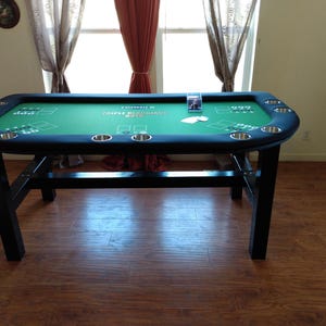May include: A rectangular poker table with a green felt surface and a black padded rim. The table has built-in cup holders and a black wooden base. The table is set in a room with hardwood floors and a window.