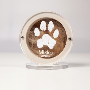May include: A circular clear acrylic memorial display frame containing a tuft of brown fur against a dark wood background. A white paw print graphic is centered above the white text Mikko and the years 2017-2026. The frame sits on a clear base.