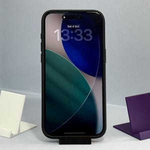 Pode incluir: Um smartphone preto com uma capa preta exibe a hora e a data. O telefone é apoiado por um suporte preto. Dois suportes adicionais, um branco e um roxo, estão em ambos os lados.