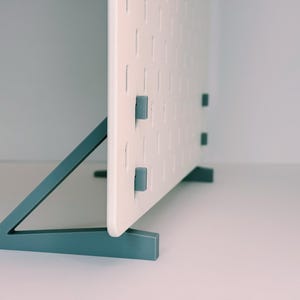 May include: A white rectangular pegboard panel featuring a grid of vertical slots, supported by two teal-coloured triangular stands. Small teal clips are inserted into the slots on the side of the board, providing structural stability and attachment points.