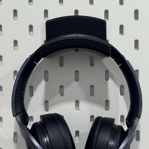 Puede incluir: Auriculares circumaurales azul oscuro colgados de un soporte negro fijado a una placa perforada blanca. Los auriculares tienen auriculares grandes y una diadema acolchada. La placa perforada tiene agujeros ovalados espaciados.