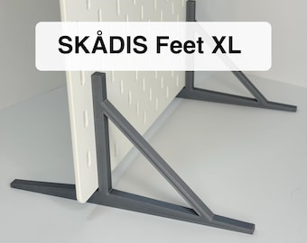 May include: Two dark gray triangular support feet holding a white pegboard upright. The feet feature a wide base and an angled brace for stability. The text SKÅDIS Feet XL is printed in black on a white rectangular background overlaying the image.