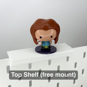 May include: A brown-haired figurine holding a small cup stands on a white shelf. The shelf is mounted on a white pegboard. The figurine has a blue and green outfit and a purple base. The text "Top Shelf (free mount)" is at the bottom.