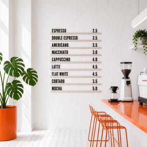 Puede incluir: Interior de una cafetería moderna con una pared blanca que muestra un menú con precios de café. Una barra naranja y taburetes a juego están en primer plano. También se ven una gran planta en maceta y equipo para hacer café.