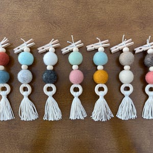 May include: A collection of eight decorative felt ball garlands. Each garland features a wooden clothespin, two felt balls, a wooden bead, a wooden ring, and a white tassel. The felt balls are in various colors.
